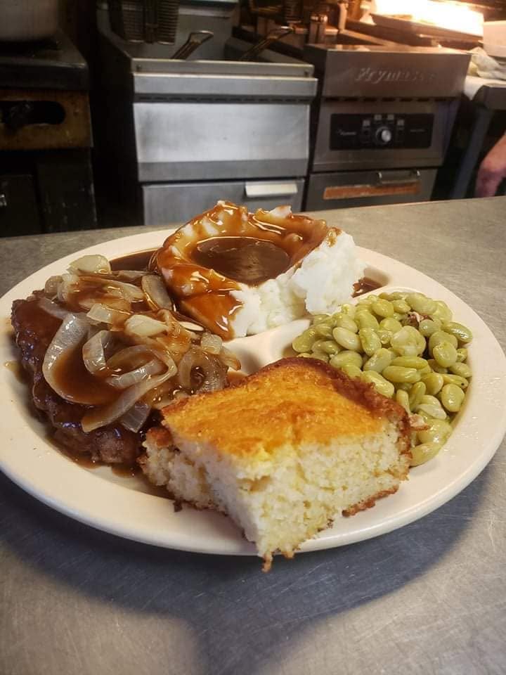 A plate of hamburger steak with gravy and onions, mashed potatoes, green beans and cornbread