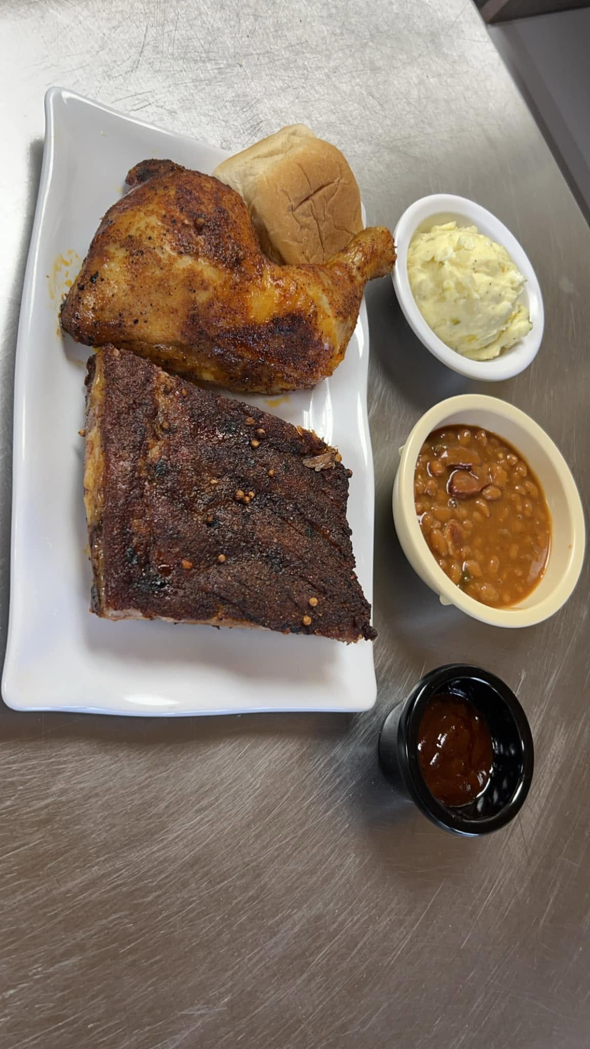 A plate of roasted chicken, a rib, potato salad, baked beans and a roll