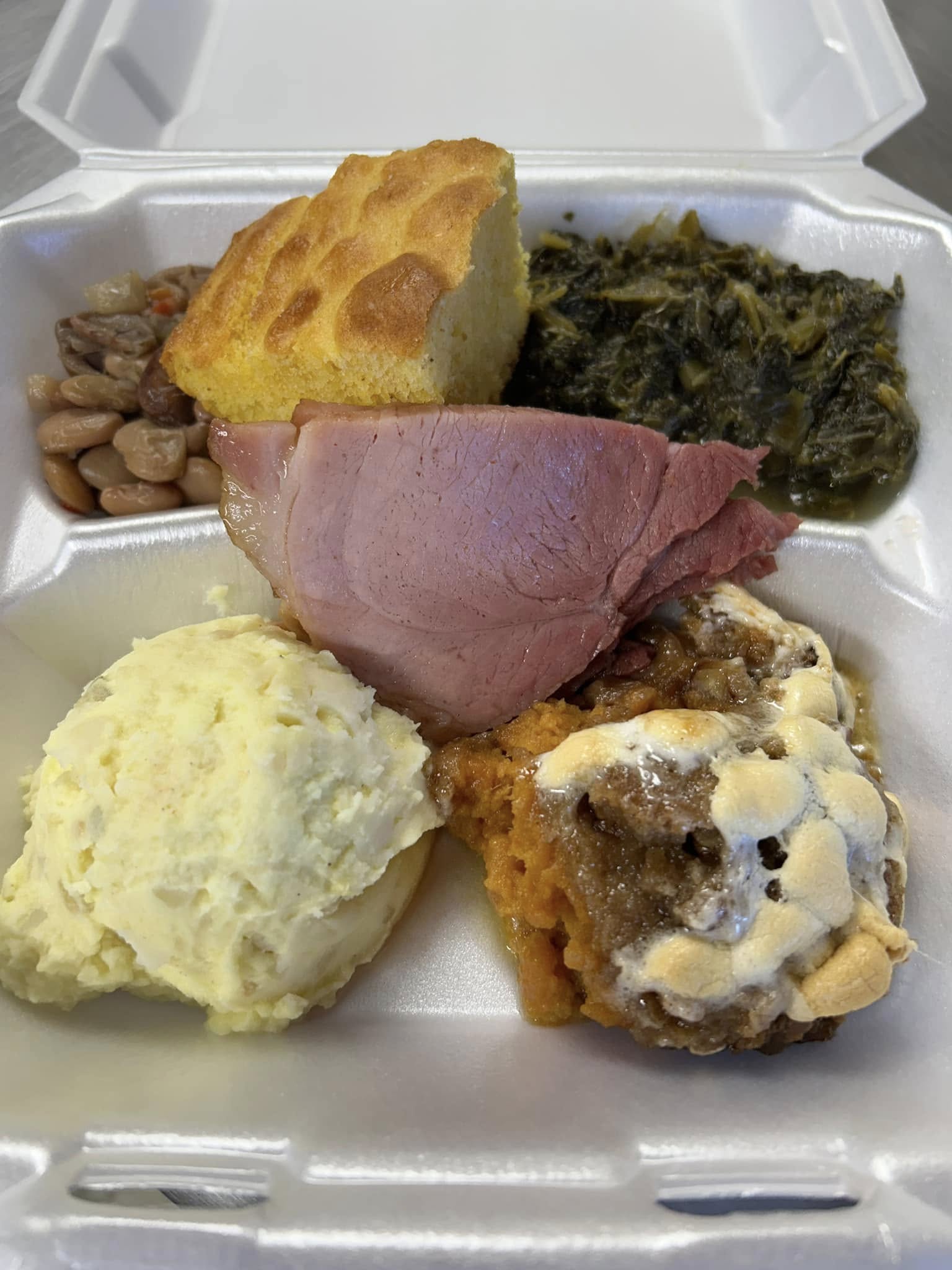 A home-style takeout plate with cornbread, greens, beans, sliced meat and mashed potatoes